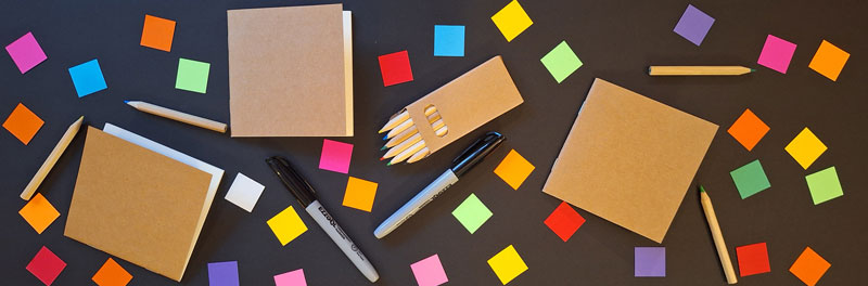 Picture of the arts supplies available for the project, small brown paper notebooks, one inch squares of colored paper, colored pencils and black markers.