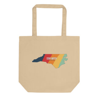 Beige colored totebag with a NC state shape with five vertical stripes - blue, red, orange, yellow and light green. Text in the center says "Stay Artsy"
