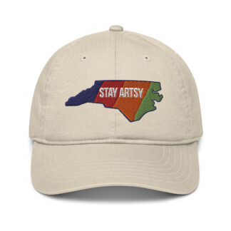a beige baseball cap embroidered with a NC state shape that has five vertical stripes - blue, red, orange, yellow and light green. Text in the center says "Stay Artsy"