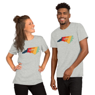 Picture of a man and a woman wearing grey heather t-shirt printed with NC state shape that has five vertical stripes - blue, red, orange, yellow and light green. Text in the center says "Stay Artsy".