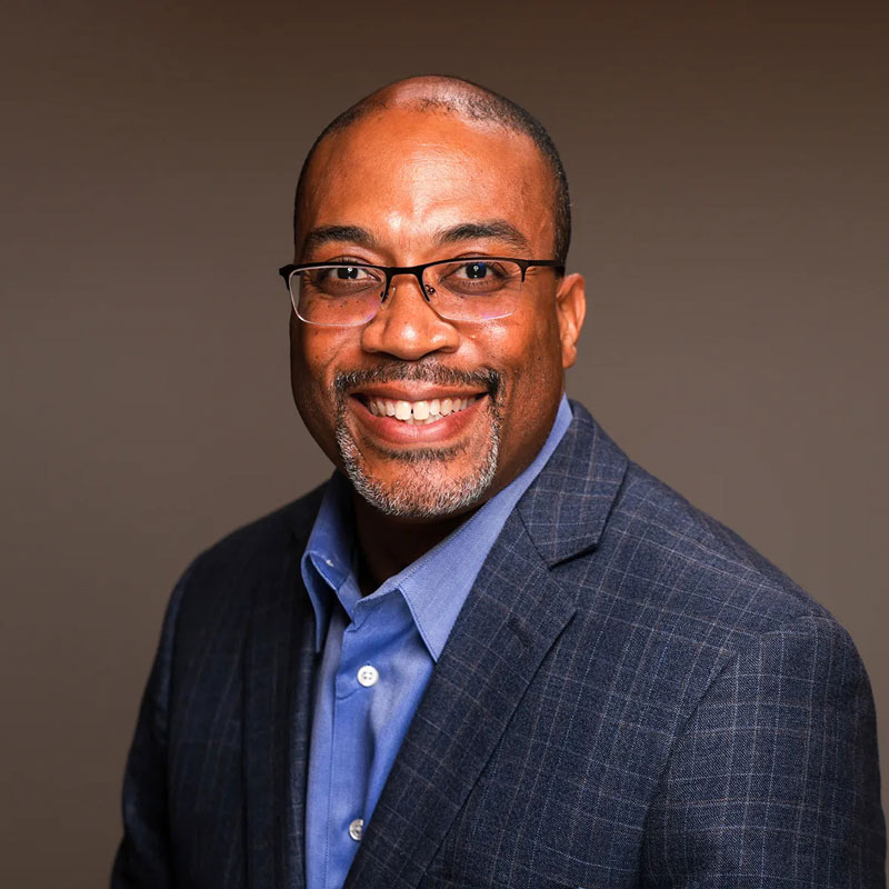 Picture of Charles Thomas, a black man with close cut hair and beard. He is wearing glasses, an open collar blue shirt and a grey suit jacket.
