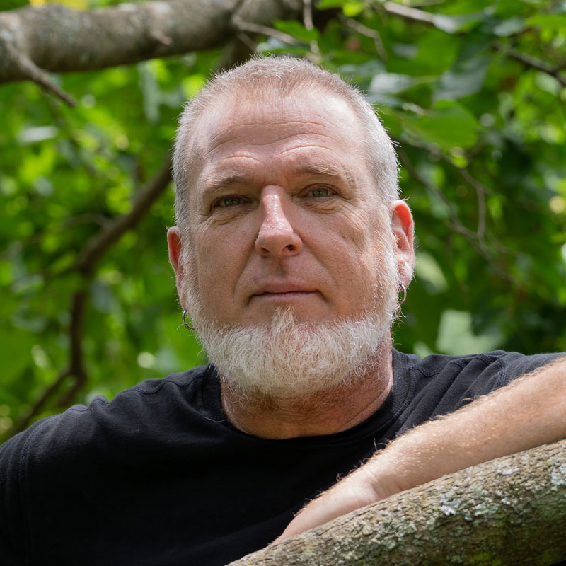 Picture of Donovan Zimmerman, a white man with short grey hair and beard. He is outside with tree leaves and brnaches behind him.
