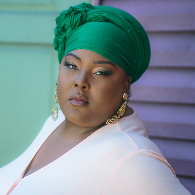 Picture of Hannah Hassan, a black woman wearing a light pink blouse. Her hair is wrapped with a green scarf and she is wearing dangling gold earings.